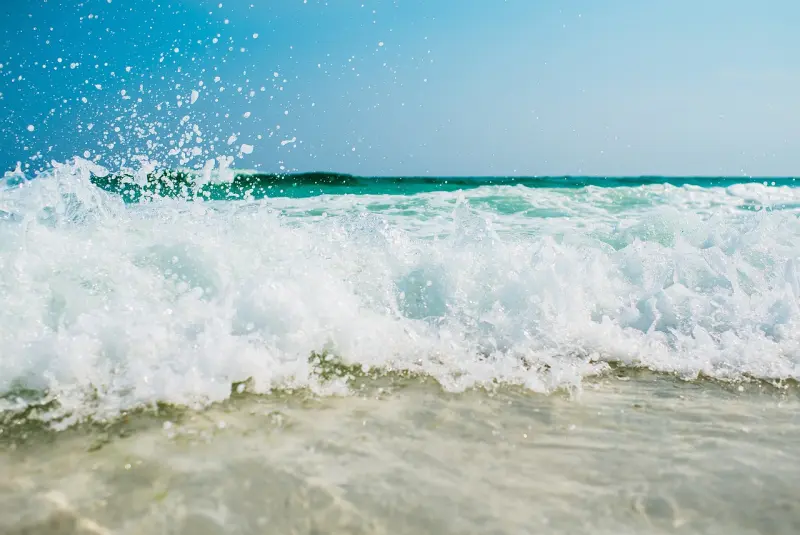 Lebendiges Bild: eine Welle bricht am Sandstrand, während Wassertropfen in der Luft tanzen.