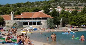 Besucher genießen den sonnigen Strand mit aufblasbaren Spielzeugen im Wasser, mit Blick auf die Steinfassade des Resorts im Hintergrund in Jezera, Murter
