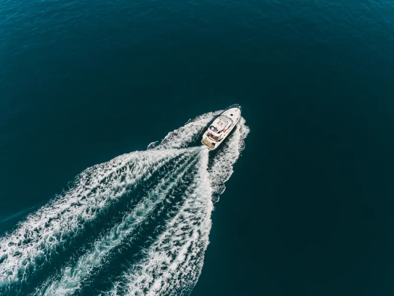 Pogled iz zraka hitrega čolna, ki ustvarja peno za seboj v globokem modrem morju, vzbujajoč občutek pustolovščine in sprostitve.