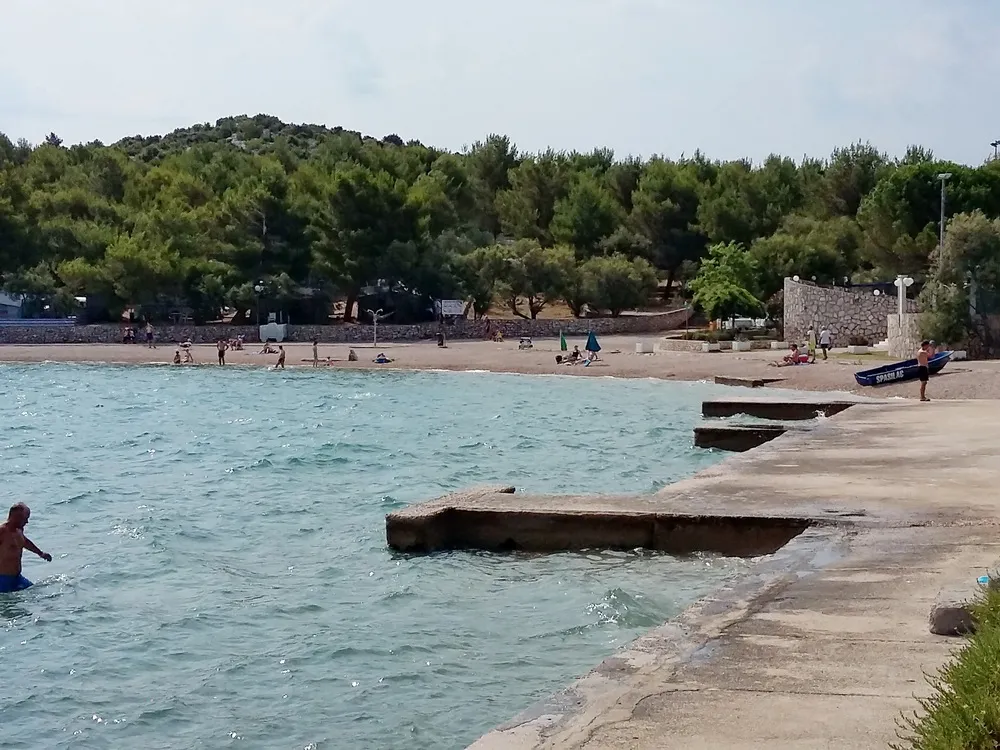 Ruhiger Strand mit Badegästen und Betonpfeilern, die in das glitzernde blaue Wasser von Jezera führen, mit üppigem Grün im Hintergrund.