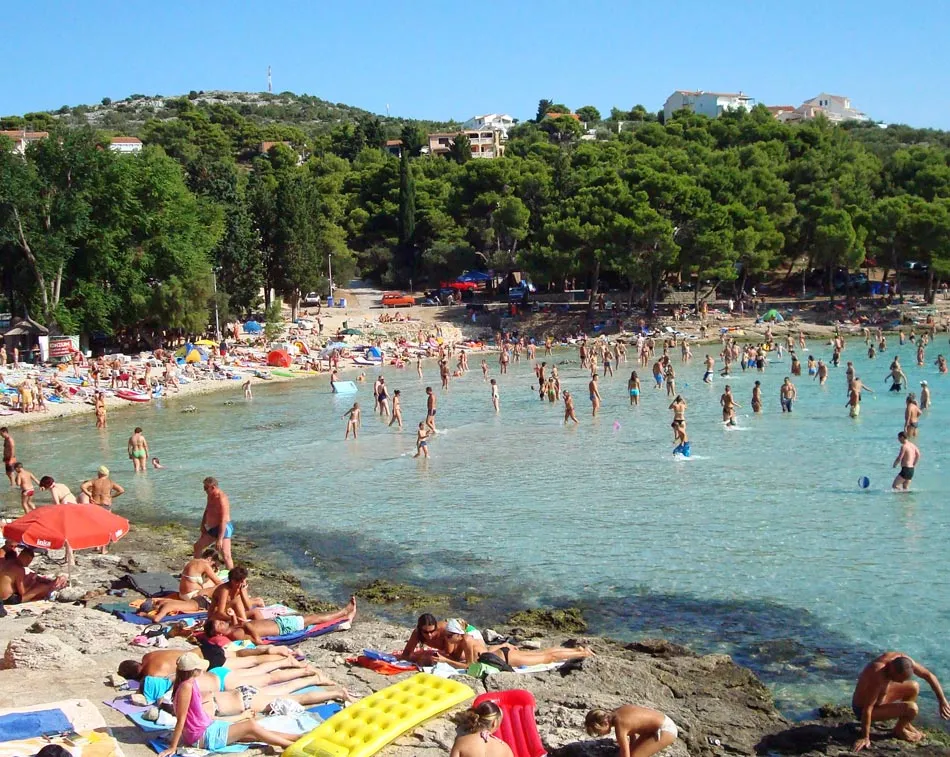 Busy summer day at a beach in Jezera, Murter with vacationers swimming, sunbathing, and enjoying the clear turquoise waters.