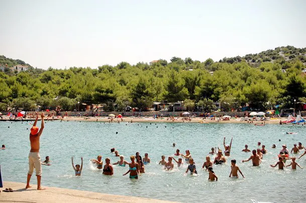 Summer day at Jezera holiday resort with tourists enjoying swimming and sunbathing on the pebbled beach with lush green trees in the background