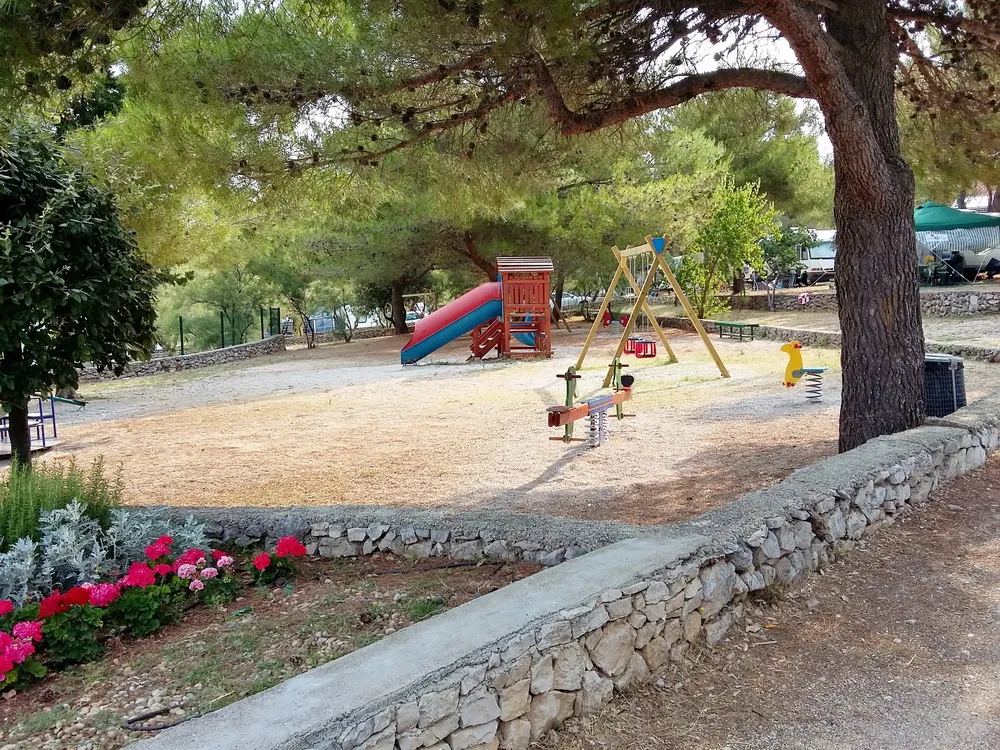 Children's playground in Jezera holiday resort featuring swings, a slide, and a seesaw on a sandy area with surrounding pine trees and colorful flowers
