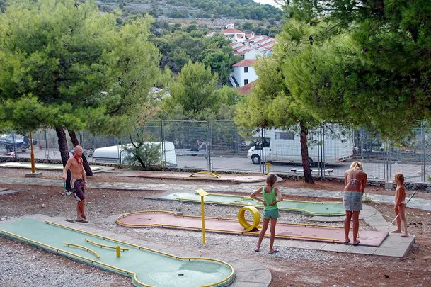 Family playing mini-golf at a resort course surrounded by pine trees with a glimpse of mobile homes and a serene holiday park setting in the background.