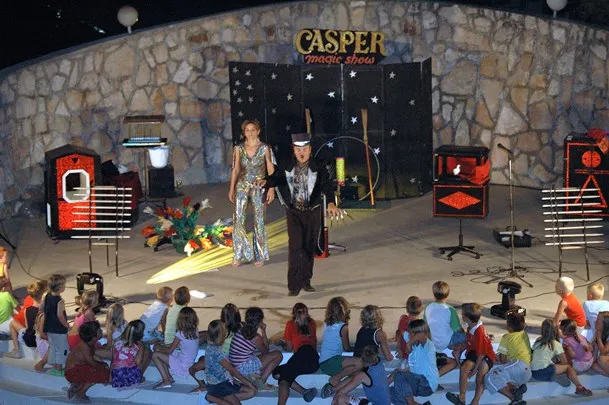 An outdoor magic show performance captivates a young audience seated in a semi-circle at night. The magician, dressed in classic attire with a top hat, and an assistant in a sparkly costume, are on stage decorated with a 'Casper Magic Show' backdrop, surrounded by colorful props and lights, creating an enchanting atmosphere.