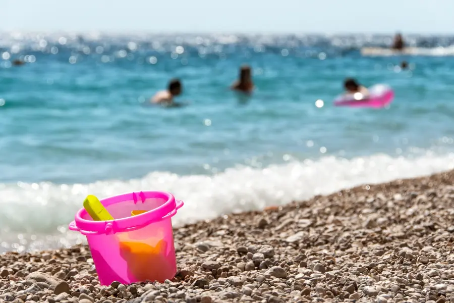Bunter Strandeimer mit Schaufel am Kieselufer, mit verschwommenen Badegästen und glitzerndem Meer im Hintergrund, ein Symbol für sommerlichen Strandspaß.
