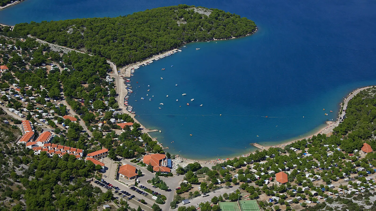 Luftaufnahme des Ferienresorts Jezera in Murter mit einem halbmondförmigen Strand, gesäumt von Booten, Mobilheimen im Grünen und klarem, blauen Wasser.