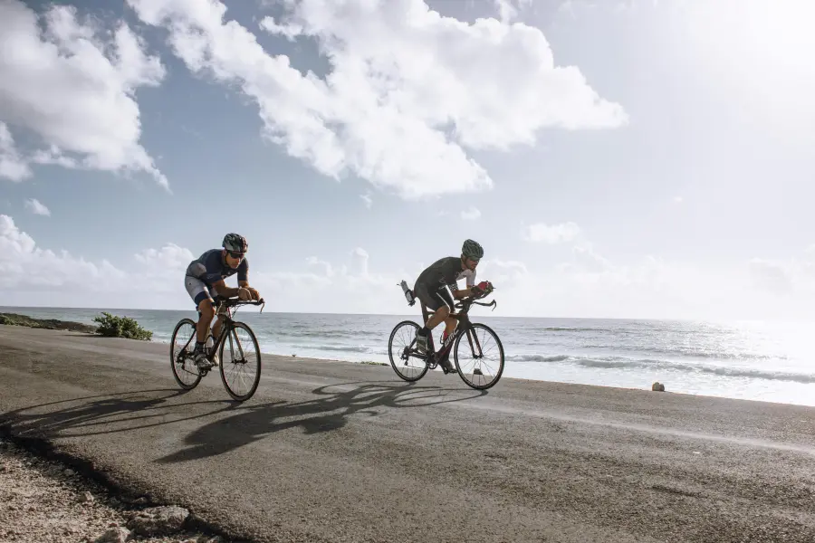 Two cyclists in racing attire riding on a coastal road beside the sea, with a sunny sky above and waves crashing in the background on Murter Island.