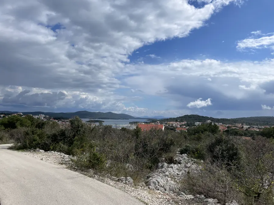 Panoramablick von einer Bergstraße aus auf die Stadt Jezera, das ruhige Meer und die sanften Hügel der Insel Murter unter einem dramatischen, wolkenverhangenen Himmel.