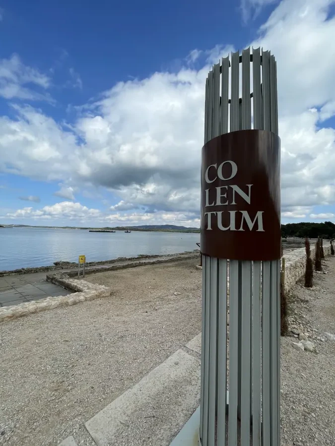 Moderna umjetnička instalacija s natpisom "COLENTUM" na povijesnom lokalitetu na otoku Murteru, s pogledom na more i dramatične oblake u pozadini.