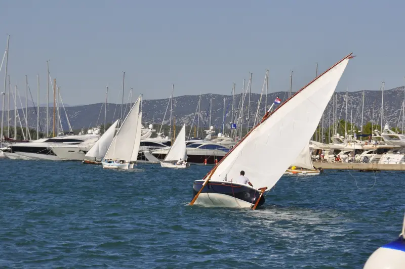 Traditional sailboat tilting in the wind with white sails unfurled, competing in a regatta, with a marina full of yachts in the background.