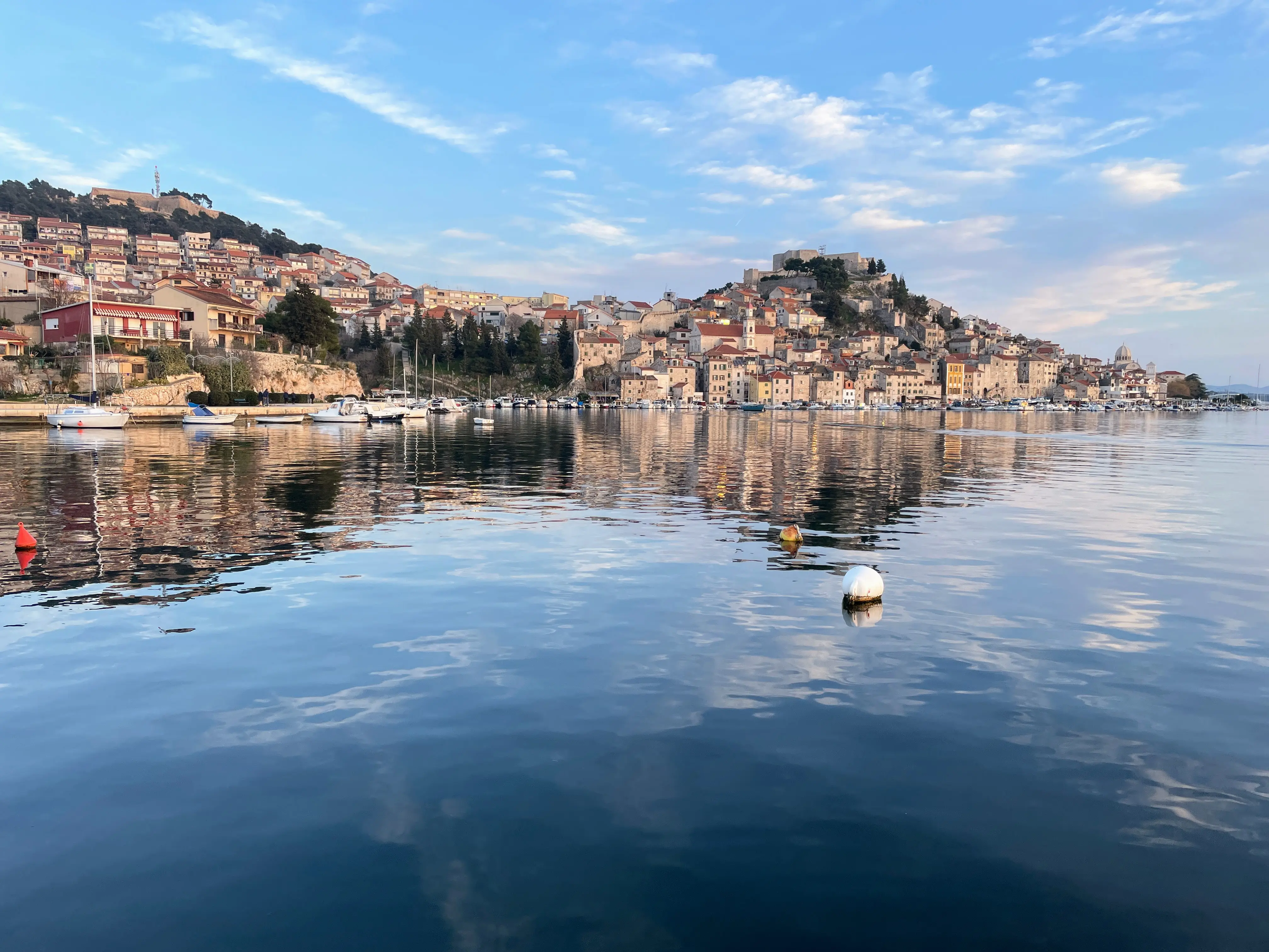 Ruhiger Hafen mit vertäuten Booten, die sich in der Abenddämmerung im Wasser spiegeln, mit Blick auf die Reihenhäuser der charmanten Stadt Šibenik