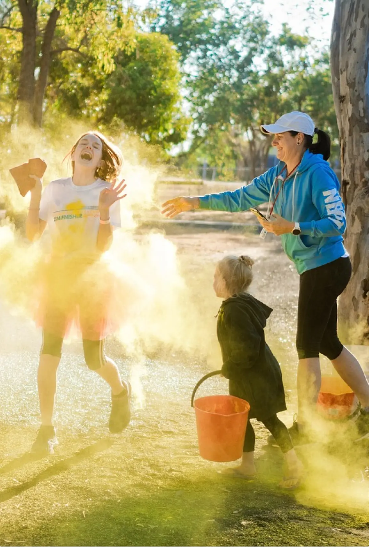 Otroci in odrasli se zabavajo med dogodkom barvnega teka, pri čemer se v zrak meče rumen prah, ki ustvarja živahen oblak