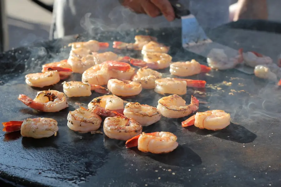 Juicy shrimps grilling on a hot plate, with steam rising, being turned by a chef's spatula, capturing the essence of outdoor cooking.