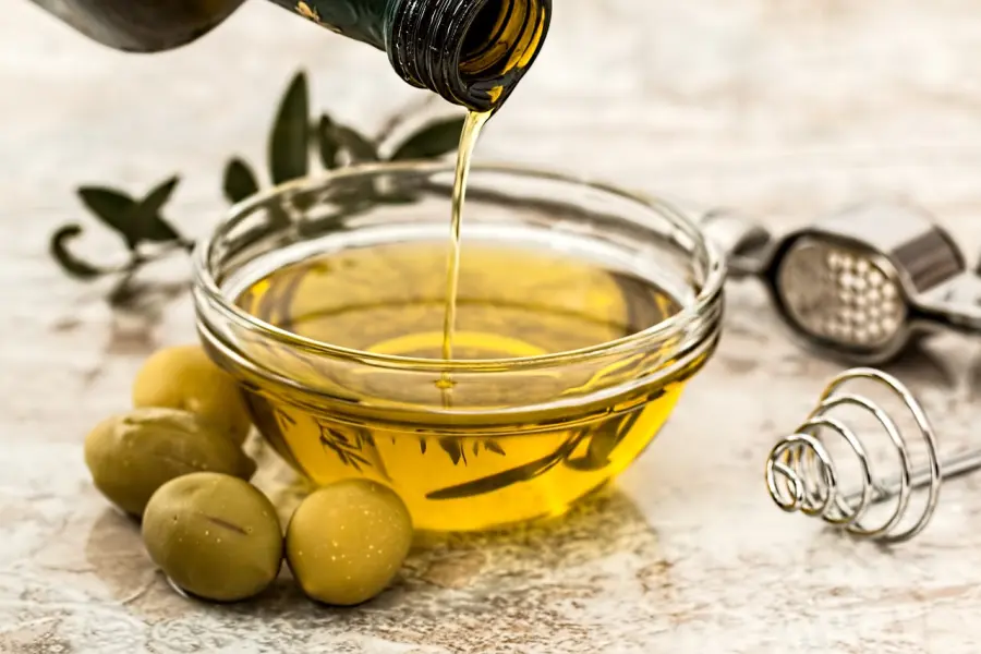 Extra virgin olive oil being poured into a glass bowl, accompanied by fresh olives, olive branches, and kitchen utensils, symbolizing Mediterranean cuisine.