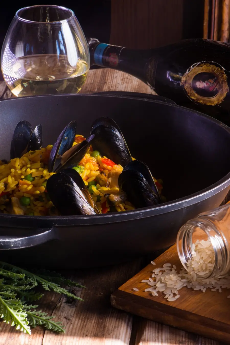 A sumptuous dish of mussels served over seasoned rice in a black pan, accompanied by a glass of white wine and a bottle in the dim ambiance, invoking a cozy dining atmosphere