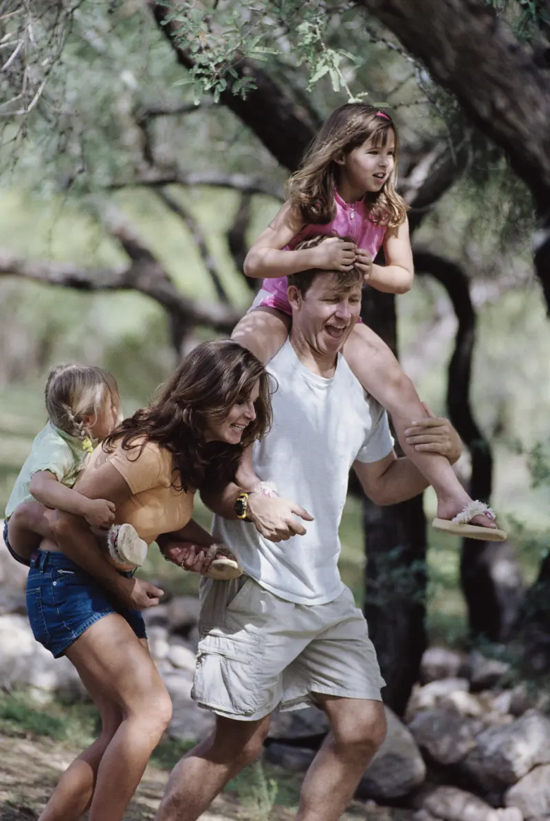 Family on a nature hike, with children riding piggyback on their parents, laughing and enjoying the outdoor adventure together
