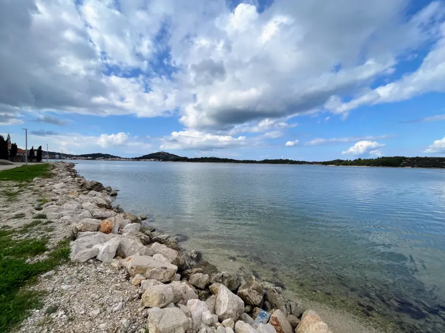 Ruhiges Wasser der Adria, umrahmt von einer felsigen Küstenlinie und mit Blick auf die Hügel der Insel Murter unter einem mit Wolken bedeckten Himmel.