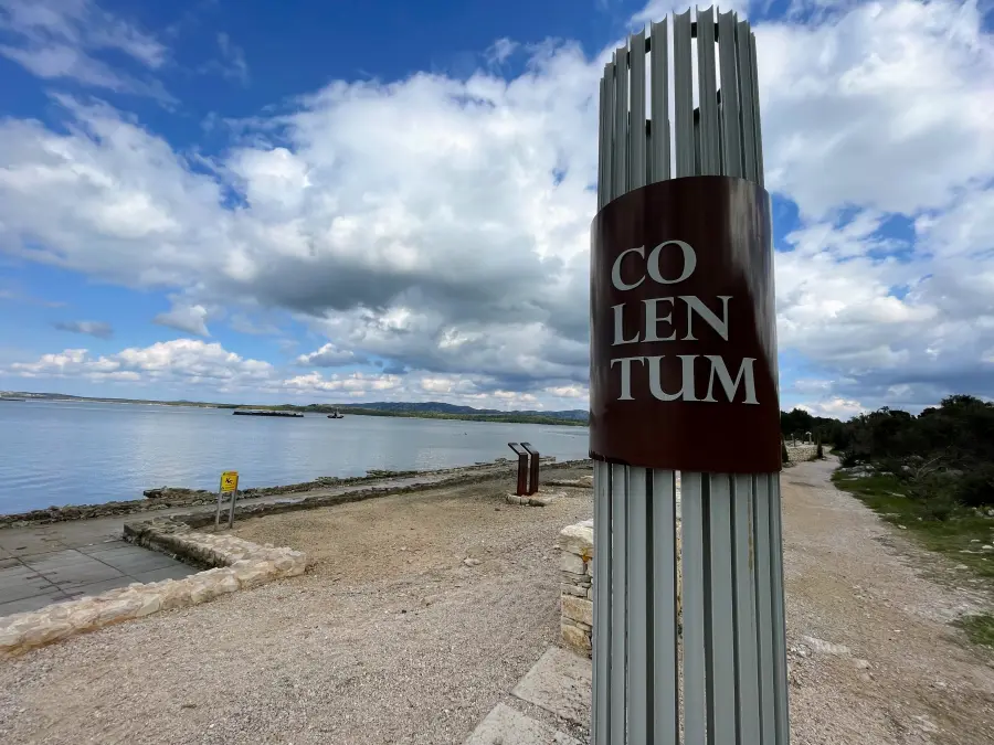 Schild mit der Aufschrift „CO LEN TUM“ auf einem Steinpfosten neben einem Strand. Der Text ist wahrscheinlich kroatisch und steht für Colentum, eine antike römische Stadt auf der Insel Murter