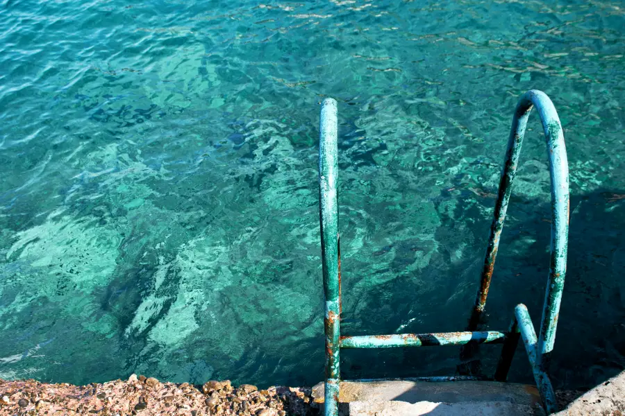 Rusty metal ladder descending into the clear, turquoise sea off the rocky shore, inviting swimmers to take a dip in the calm waters