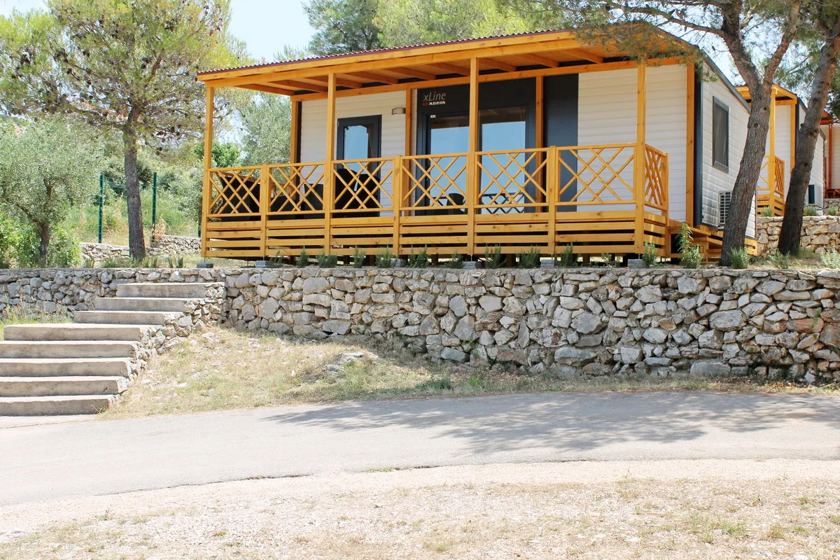 Ein charmantes Mobilheim mit einer gelben Holzterrasse, das sich über einer Steinmauer erhebt und von mediterranen Kiefern umgeben ist.