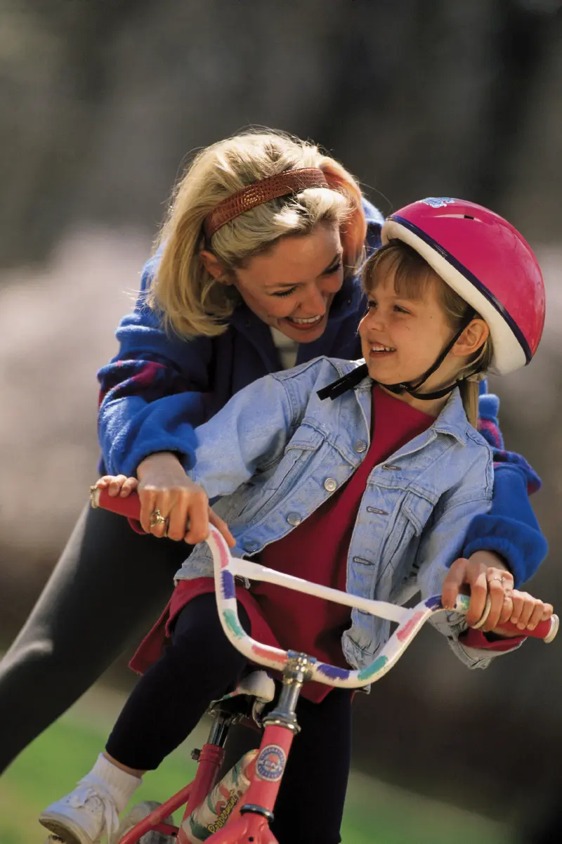 Radosten trenutek, ko mati pomaga svoji hčerki voziti kolo, pri čemer ima otrok na glavi rožnato čelado in velik nasmeh.
