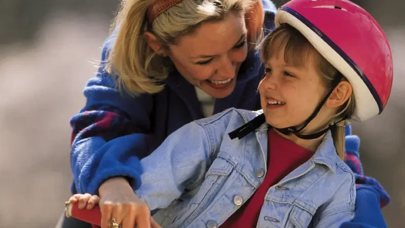 Fröhlicher Moment, als eine Mutter ihrer Tochter beim Fahrradfahren hilft, wobei das Kind einen rosa Helm trägt und breit lächelt.