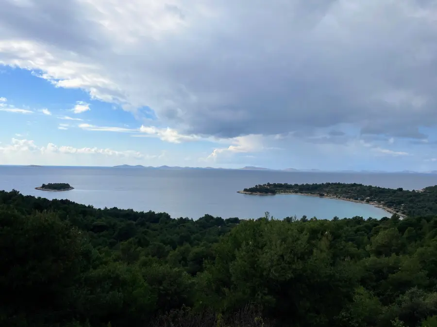 Slikoviti pogled na Jezera na Murteru s bujnim zelenilom u prvom planu, mirnim Jadranskim morem i oblačnim nebom u pozadini.